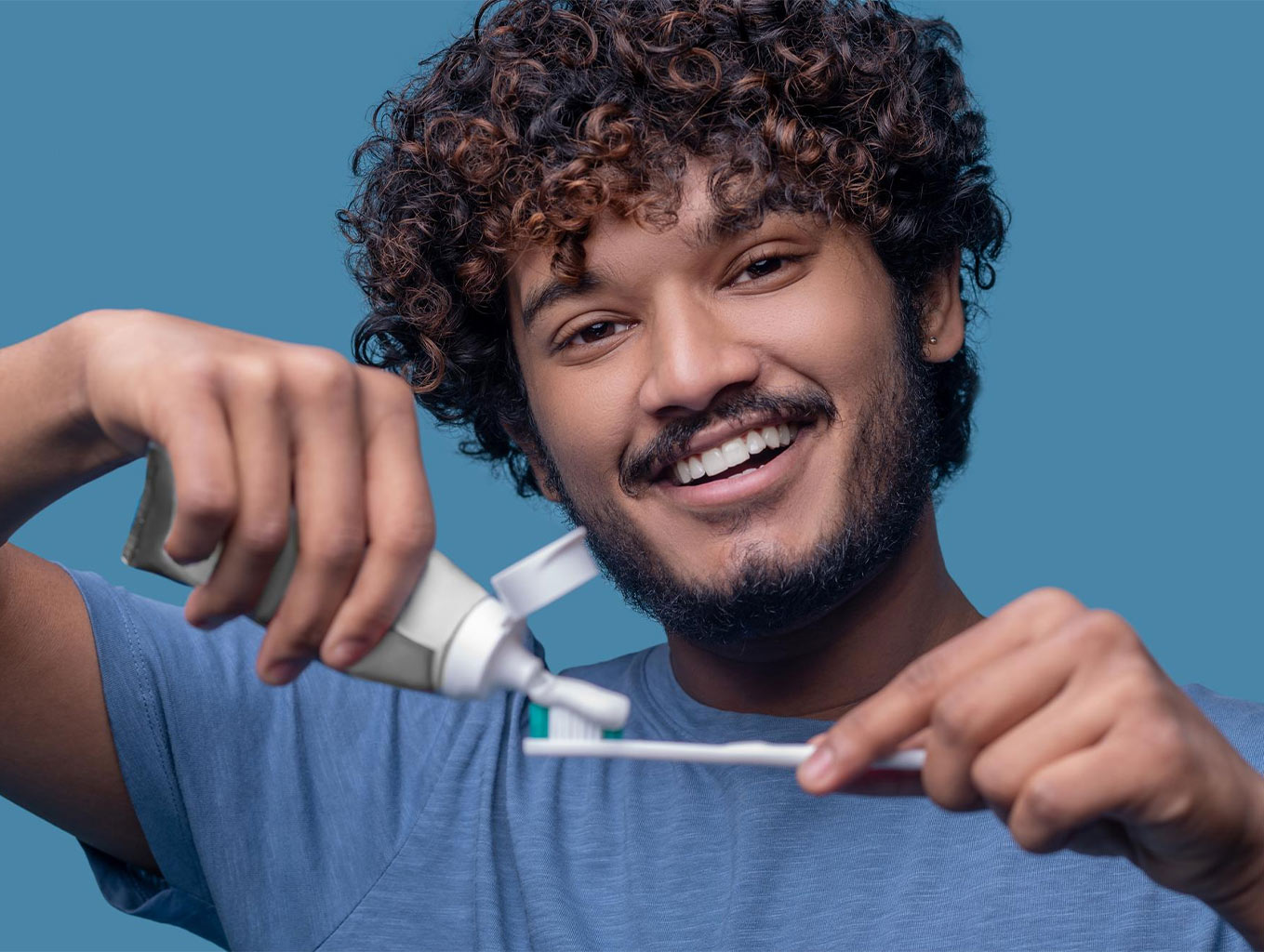 در این تصویر، یک مرد جوان با موهای فر و چهره بشاش در حال گذاشتن خمیردندان روی مسواک است. عکس حس مثبت، انرژی و سلامت را منتقل میکند و برای محتواهای آموزشی درباره مسواک زدن، معرفی خمیردندان یا تبلیغات بهداشتی بسیار مناسب است.